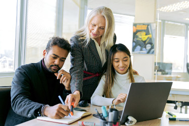 A man and a woman are seated in an office, looking at a computer, while a woman wearing a suit stands behind them and leans in to look at their work. 