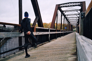 A man wearing exercise clothing stretches his legs as he prepares to run across a bridge.