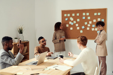 A group of coworkers is in a meeting and discussing sticky notes attached to a corkboard at the front of the room.